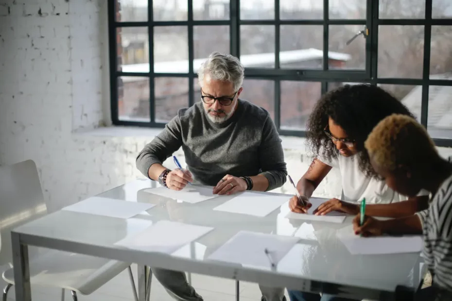 three people writing while sat at glass table in industrial room with white brick walls